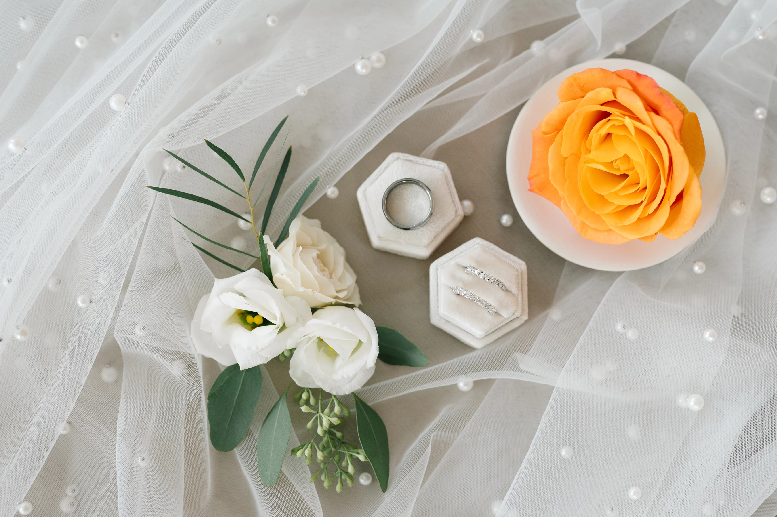 Lace Fabric With Pearls, White Flowers, And An Orange Rose Beside Ring Boxes With Silver Rings